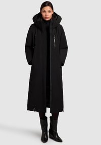 Long black puffer coat with a hood, front zipper, and pockets. Smooth texture, insulated design, and a straight silhouette. Black boots below.
