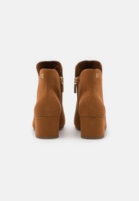Botas de tobillo de ante marrón con tacones de bloque, cremalleras doradas laterales y pequeños logotipos redondos dorados en la parte trasera externa de cada bota.