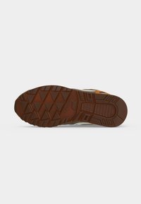 Semelle de chaussure de sport marron avec des motifs de bande de roulement géométriques et le nom de la marque embossé, présentée sur fond blanc uni.