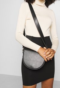 Sac bandoulière synthétique noir de forme arrondie, avec une sangle ajustable et une texture subtile. Porté avec une jupe noire ajustée et un haut beige.