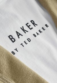 Valge t-särk, millel on must kiri "BAKER" ja "BY TED BAKER". Kaunistatud helepruuni kangast aktsendiga ümber kaela. Sile tekstuur.