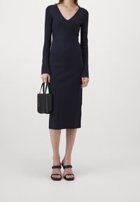 Robe longue bleu marine à manches longues avec un col en V, présentant une silhouette ajustée et des détails de couture en losange. Associée à un petit sac à main noir et des sandales.