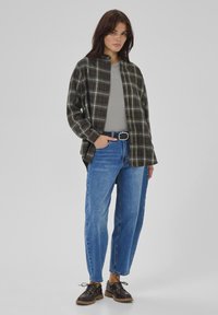 Kvinde iført en grå skjorte, en overdimensioneret mørkternet jakke, blå højtaljede jeans, sort bælte og sorte snøresko, stående med den ene hånd i lommen.