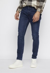 Jeans skinny de mezclilla azul oscuro con una textura suave, combinados con zapatillas altas blancas. La camisa presenta un patrón de cuadros en beige y marrón.