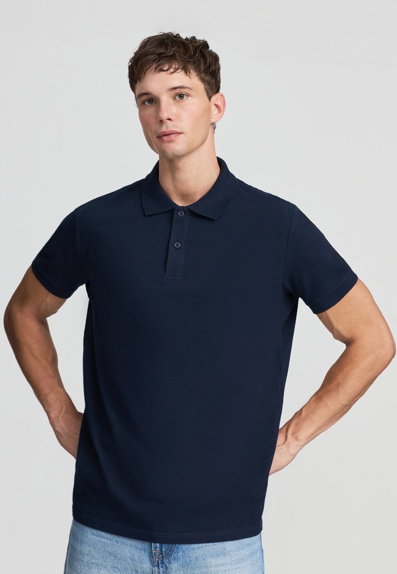 Giovane uomo in piedi con le mani sui fianchi, che indossa una polo a maniche corte blu scuro e jeans azzurri, su uno sfondo neutro.