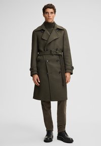 Manteau trench en laine double boutonnage vert olive avec une ceinture, grands revers et détails de boutons, associé à un pantalon olive et des bottes noires.