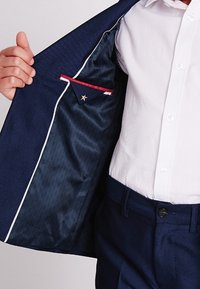 Granatowa marynarka o teksturowanej powierzchni, z białą podszewką i wewnętrzną kieszenią z czerwonym akcentem, zdobioną detalem w kształcie gwiazdy. Nosi się ją na białej koszuli.