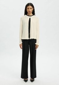 Woman with long dark hair wearing cream cardigan over black top, black wide-leg trousers, and black pointed shoes standing on a plain background.