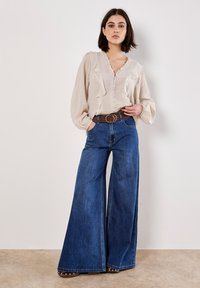 Krämfärgad blus med volanger, knapplist framtill; kombinerad med vida blå jeans, prydda med ett brunt bälte och detaljer i silver.