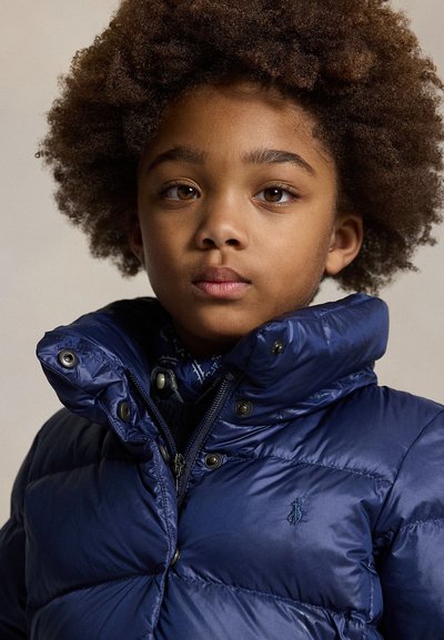 Bambino con capelli ricci che indossa una giacca imbottita blu con colletto rialzato, che guarda dritto davanti a sé con espressione neutra su sfondo semplice.