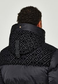 Mand vender ryggen til iført en sort puffjakke med geometrisk mønster på huen og øvre ryg, der er udstyret med en sort stofsløjfe og trykknap.