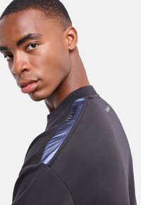 Sudadera negra con cuello redondo, acentos texturizados en los hombros en azul y un acabado suave con detalle de etiqueta en la parte superior de la espalda.