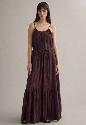 Vrouw draagt een mouwloze, donkergroene geplooide maxi-jurk met dunne bandjes en een gestrikte tailleband, staand tegen een effen achtergrond.