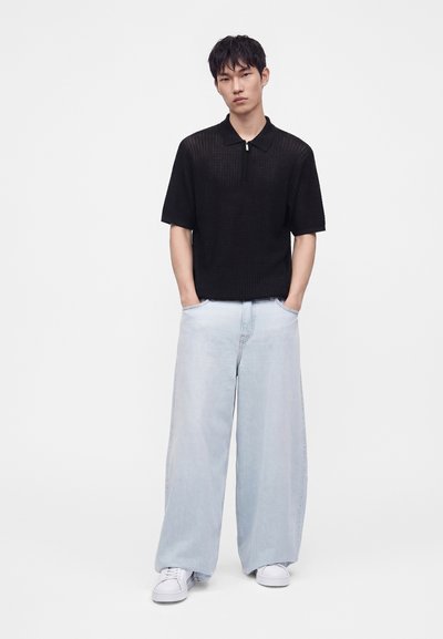 Homme portant une chemise noire à manches courtes zippée, un jean large bleu clair et des baskets blanches, debout les mains dans les poches sur fond blanc uni.