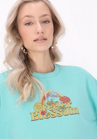 Türkiz kapucnis pulóver, kerek nyakkivágással és „Feelin' Blossom” virágos grafikával. A modellen színes karika fülbevalók és göndör haj látható.