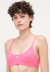 Rožinė sportinė liemenėlė su plonais dirželiais, pagaminta iš tamprios medžiagos. Turi išlenktą iškirpimą ir mažą baltą "Nike" logotipą priekyje.