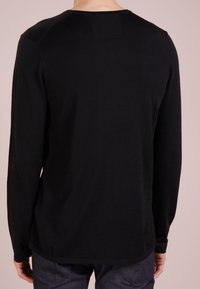 Persona che indossa una maglietta nera a maniche lunghe e jeans scuri, mostrata di spalle su uno sfondo neutro.