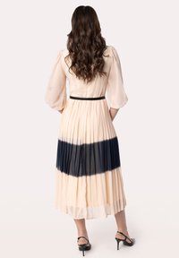 Abito plissettato in pesco chiaro con effetto ombre blu navy sull'orlo, con maniche trasparenti e una cintura nera in vita, abbinato a tacchi neri.