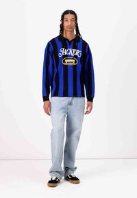 Blue and black striped sweater with a hood, featuring a logo in yellow on the front. Paired with light blue jeans and black sneakers.