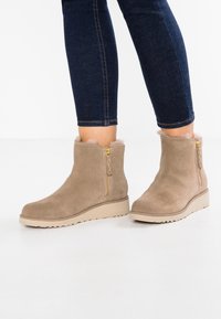 Botas de tobillo de ante marrón claro con un acabado texturizado, que presentan un cierre lateral con cremallera, un forro suave y una suela de goma beige.