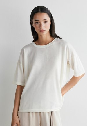 Ung kvinde med langt mørkt hår iført løs hvid kortærmet skjorte og lysebeige bukser, stående med den ene hånd i lommen mod en ensfarvet væg.