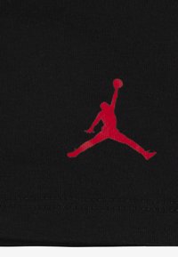 Schwarzer Stoff mit einem roten Jumpman-Logo, das eine silhouettierte Figur zeigt, die mit einem Basketball in der Hand springt. Details umfassen eine glatte Textur und Nähte.