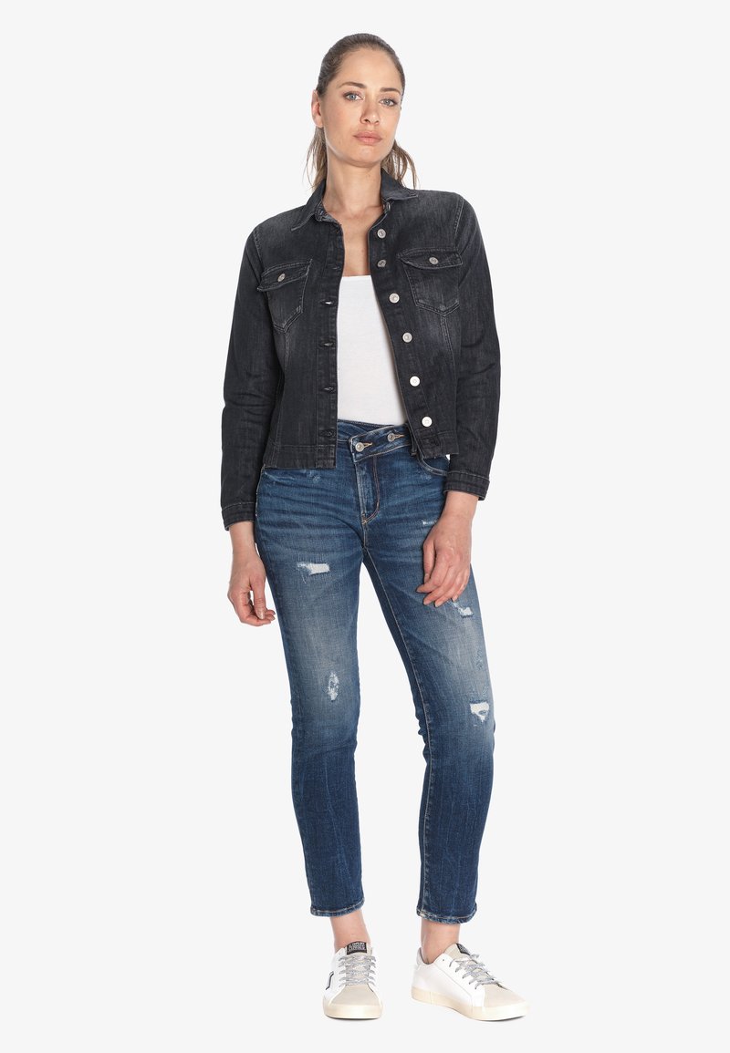 Veste en denim noir, chemise blanche, jean bleu déchiré et baskets blanches. La veste a des détails de boutons ; le jean est ajusté au niveau des chevilles.