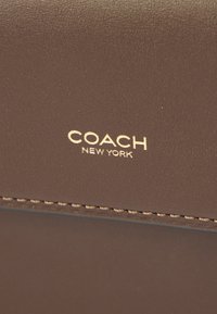 Surface en cuir marron avec logo embossé en or "COACH NEW YORK" et coutures visibles le long du bord.