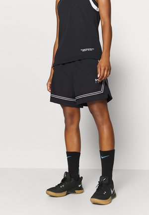 Person wearing black athletic shorts, sleeveless shirt with text, tall black socks, and black Nike sneakers standing on a white floor.