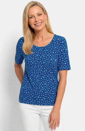 Smiling woman with long blonde hair wears a blue short-sleeve top with light blue spots and white pants, standing against a plain background.