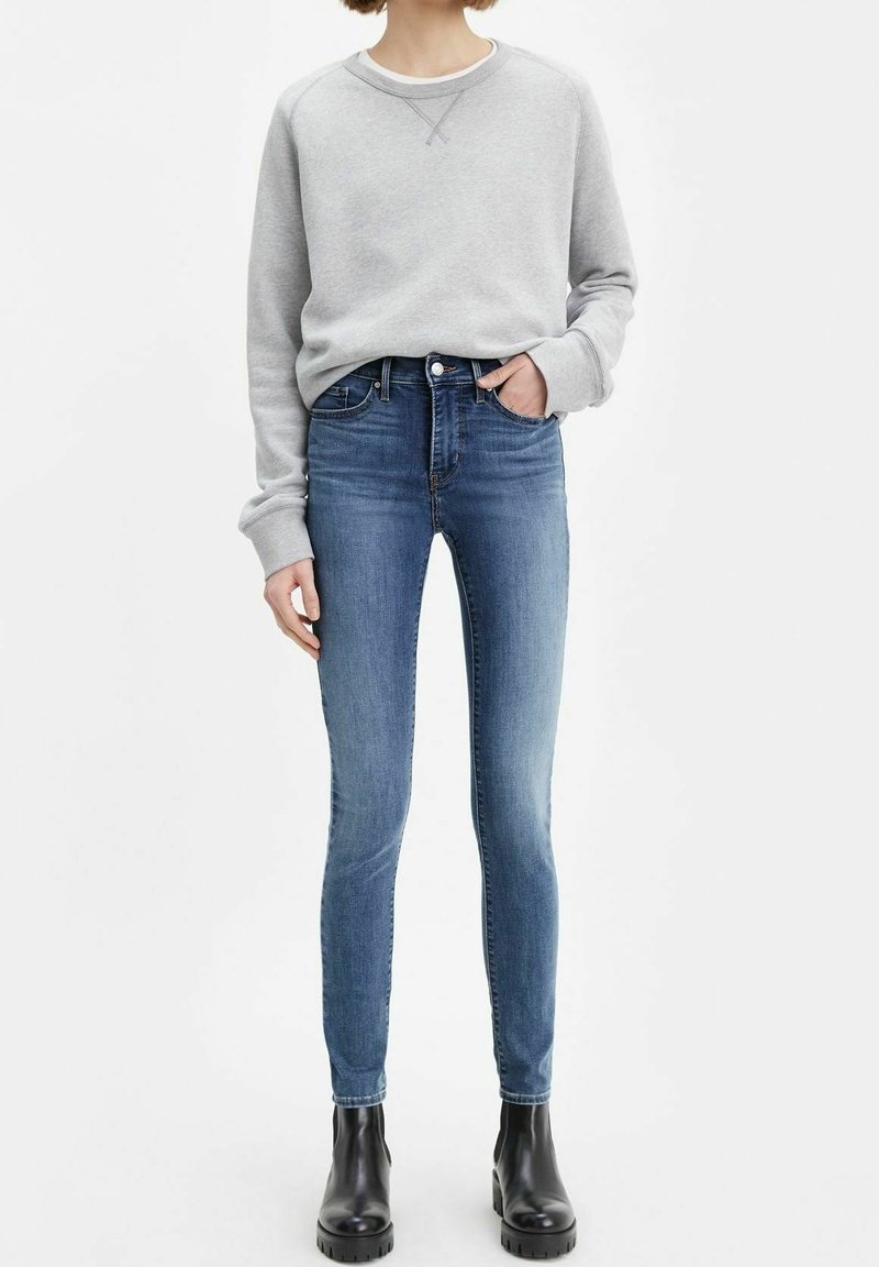 Personne portant un sweat-shirt gris clair, un jean skinny bleu et des bottines noires, debout avec une main dans la poche.