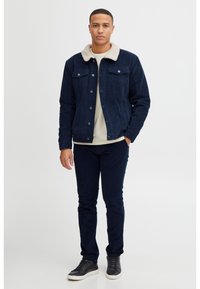 Chaqueta de pana azul marino con un cuello de piel sintética beige, bolsillos frontales y cierre de botones. Combinada con pantalones oscuros y zapatillas negras.