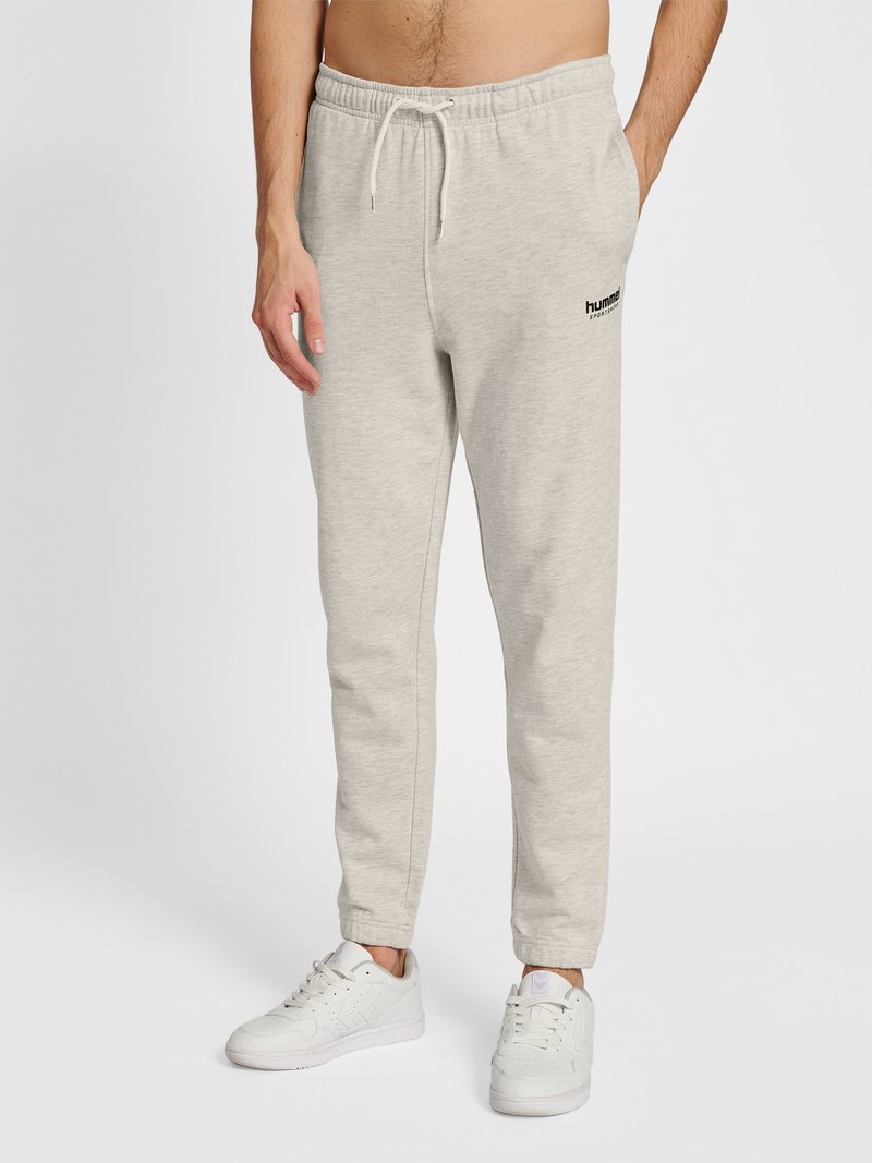 Homme portant un pantalon de jogging gris clair avec taille à cordon et des baskets blanches, debout devant un fond uni.