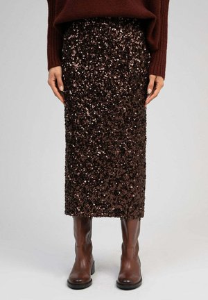Femme portant une jupe midi à sequins marron foncé, un pull en maille marron et des bottes en cuir marron montant jusqu'aux genoux, debout devant un fond uni.