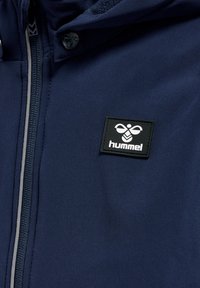 Navyblå zip-up jakke med fleeceforing, der har et sort rektangulært "Hummel" logo-patch på venstre brystområde.