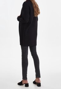 Dlouhý černý pletený cardigan s texturovaným vzorem, doplněný o tmavě šedé úzké džíny a černé mule s otevřenou špičkou, pohled zezadu.