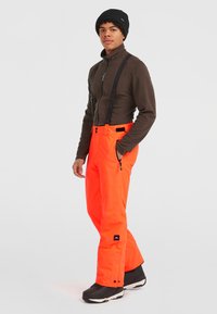 Ljusorange skidbyxor med justerbara svarta remmar, fickor med dragkedja och en figurnära design. Bärs med en brun fleecetröja och en svart mössa.