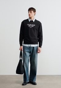 Άνδρας που φοράει μαύρο φούτερ Calvin Klein, ριγέ πουκάμισο από κάτω, μπλε τζιν, μαύρα παπούτσια, κρατάει μαύρη τσάντα tote, στέκεται μπροστά σε λευκό τοίχο.