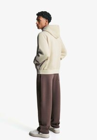 Světle béžová mikina s kapucí a volným střihem, v kombinaci s hnědými tepláky, které mají boční kapsy a jsou doplněny světle barevnými teniskami.