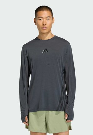 Joven con una camiseta deportiva de manga larga color gris oscuro con aberturas para el pulgar y pantalones cortos verdes claros, de pie contra un fondo liso.