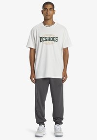 White cotton t-shirt with green and gold DC Shoes logo, paired with gray sweatpants featuring a subtle logo, and white athletic shoes.