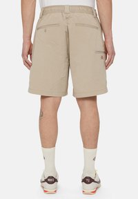Beige cargo shorts dengan sabuk elastis, dua saku belakang, dan tambalan logo dekoratif. Kain yang ringan dan berb-texture.
