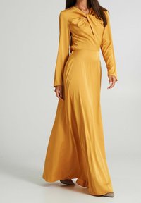 Långärmad klänning i senapsgul färg med en knutdetalj vid halsen, flödande siluett och mjuk textur. Glittrande skor synliga.