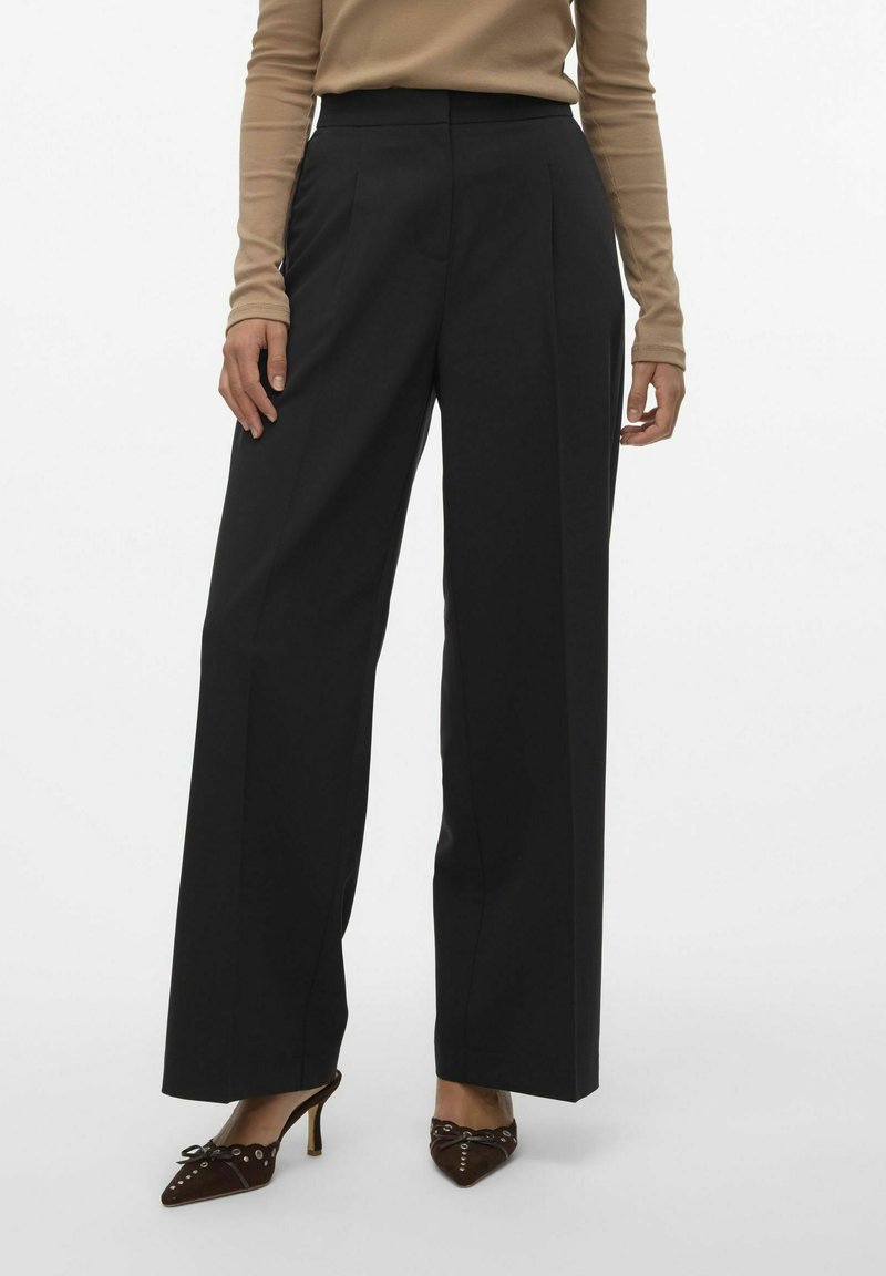 Zwarte wijde broek gemaakt van een gladde stof, met een hoge taille en plooien aan de voorkant. Draagt bij bruine puntige hakken.