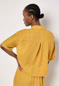 Senfgelbe kurzärmlige Bluse mit lockerer Passform, Kragen und Rückenschlitz. Leichter Stoff mit glatter Textur.