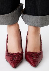 Zapatos de piel roja con punta afilada que cuentan con detalles de cordones en la parte delantera y acentos de corte, usados con jeans oscuros de denim arremangados.