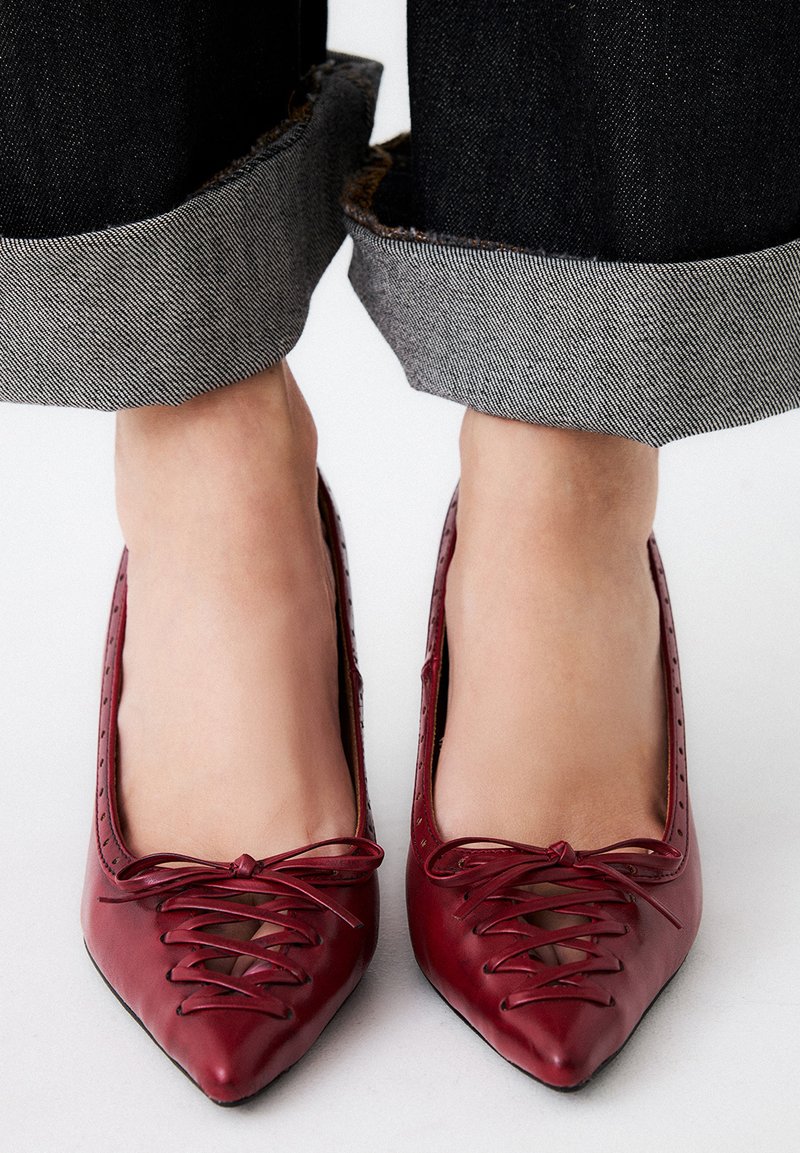 Zapatos de piel roja con punta afilada que cuentan con detalles de cordones en la parte delantera y acentos de corte, usados con jeans oscuros de denim arremangados.