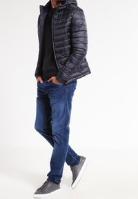 Giubbotto imbottito nero con cappuccio, tessuto testurizzato, chiusura frontale con zip. Indossato sopra un maglione nero, abbinato a jeans blu e sneakers grigie.