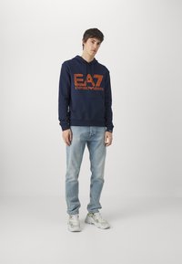 Felpa blu navy con logo arancione, in mischia di cotone, con tasca a marsupio, abbinata a jeans chiari e sneakers bianche. Texture liscia, vestibilità casual.