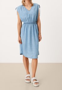 Ljusblå denimklänning med V-ringning, elastisk midja och knyt detaljer vid axlarna. Slät textur, knälängd med sidoklyftor.